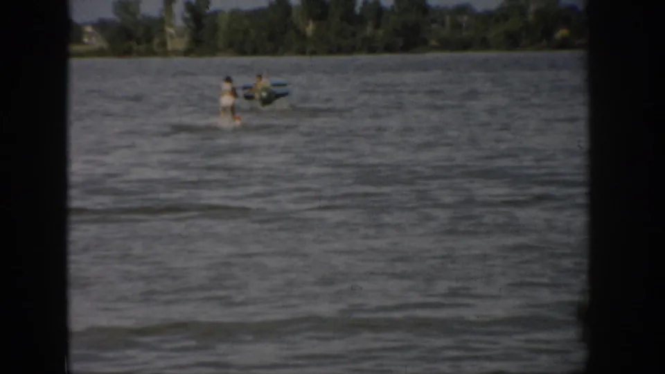 1959 Colorado home-movie frame showing a distant woman water skiing near a small motorboat on a windy lake.
