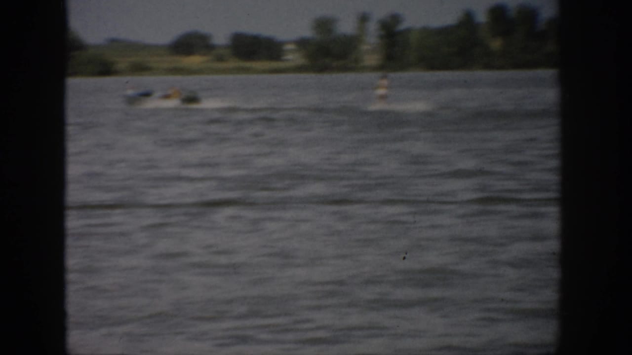 1959 Colorado home-movie view across a lake toward small boats and a tree-lined shore, seen through dark foreground framing.