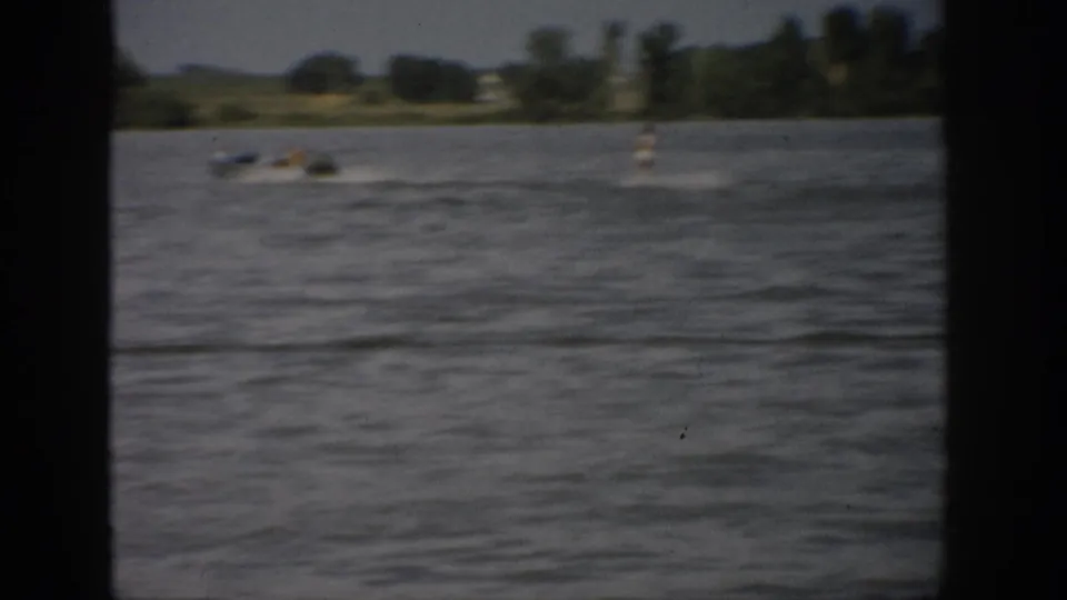 1959 Colorado home-movie view across a lake toward small boats and a tree-lined shore, seen through dark foreground framing.