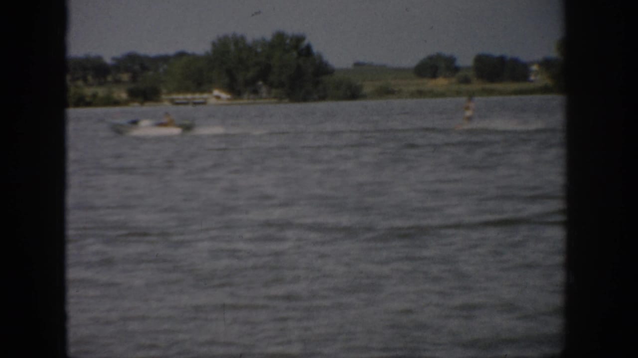 1959 Colorado lake scene showing a small motorboat on open water with trees, fields, and dark frame edges in the distance.