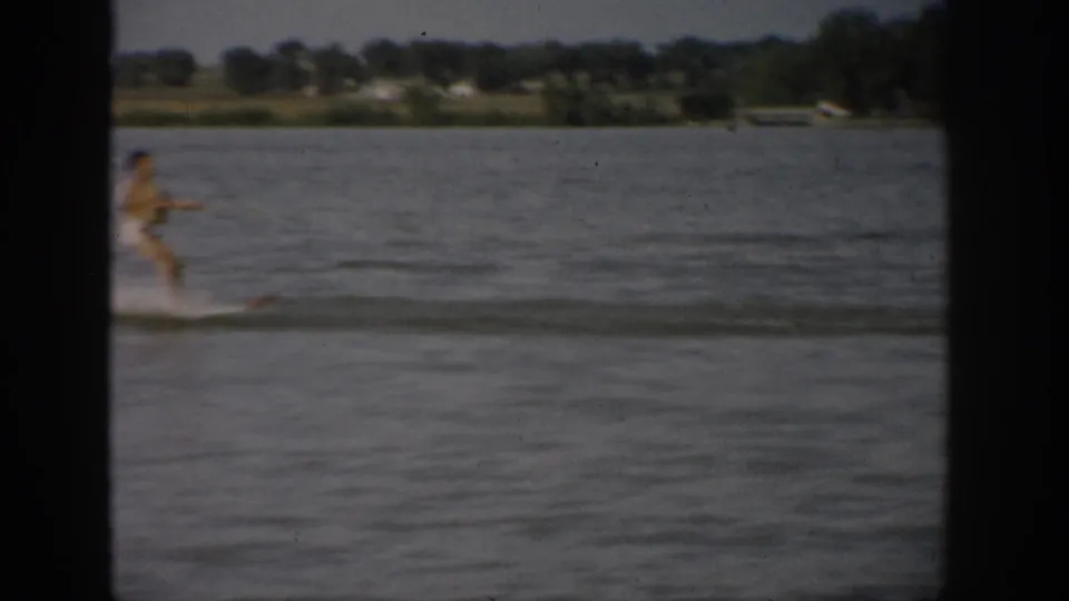 1959 Colorado home-movie frame showing a water skier gliding across a lake behind a towing boat, with fields and trees on the far shore.