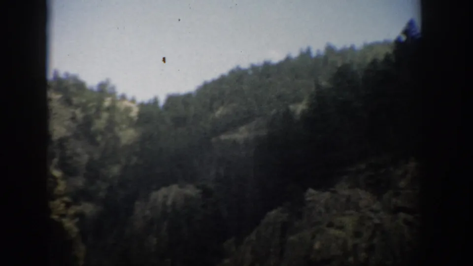 1959 Colorado home-movie frame with wooded mountain slopes, brushy foreground, and heavy edge shadow from the film gate.