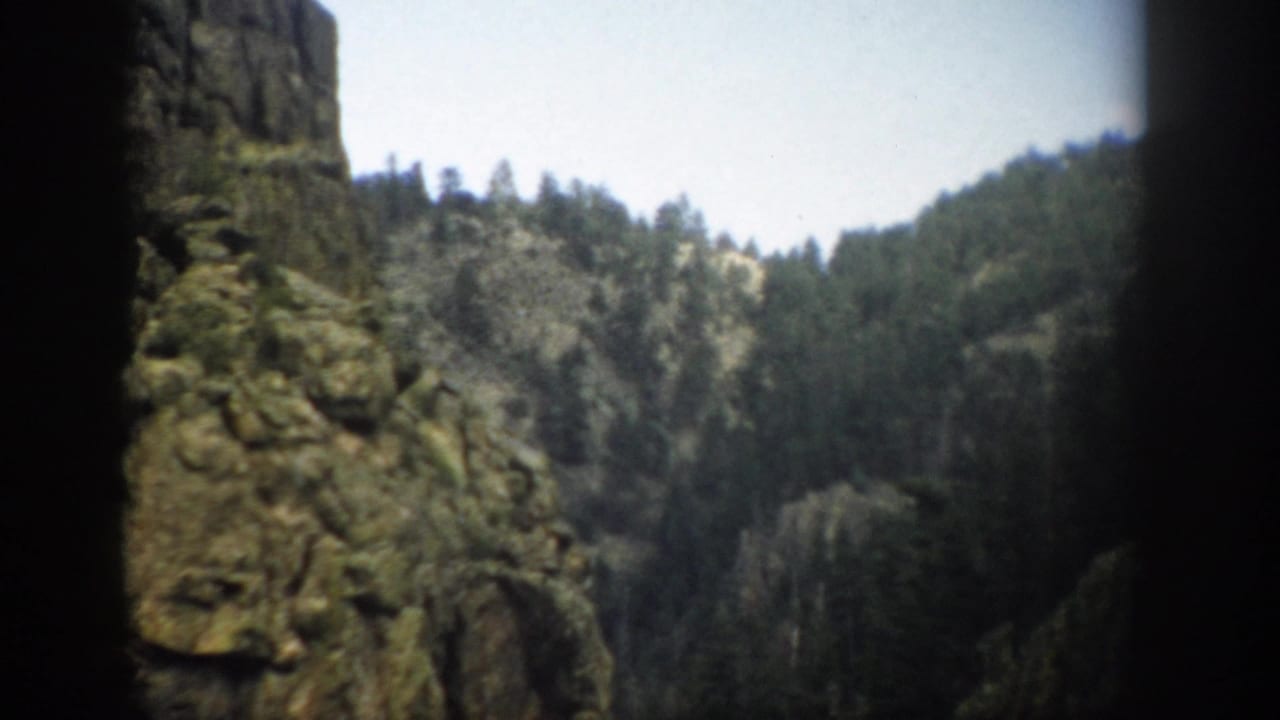 1959 Colorado 8mm frame showing a distant evergreen-covered mountain vista seen between dark foreground rock edges.
