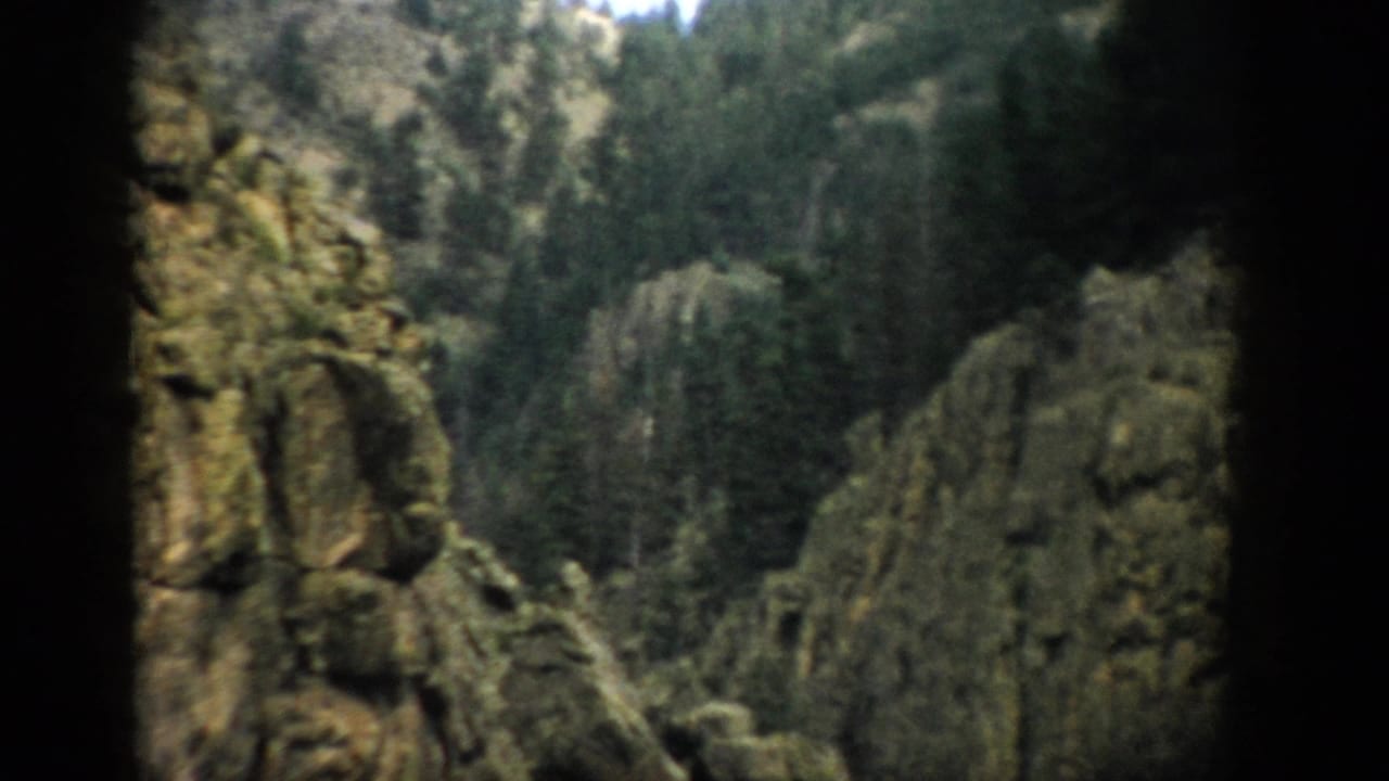1959 Colorado home-movie view of rocky cliffs and dense evergreen forest framed by shadowed foreground stone.