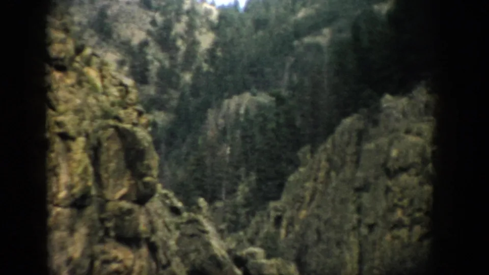 1959 Colorado home-movie view of rocky cliffs and dense evergreen forest framed by shadowed foreground stone.