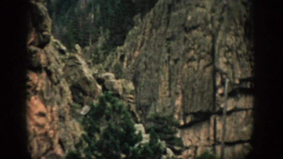 1959 Colorado home-movie frame with mossy rock walls, evergreen growth, and a dark vertical foreground edge.