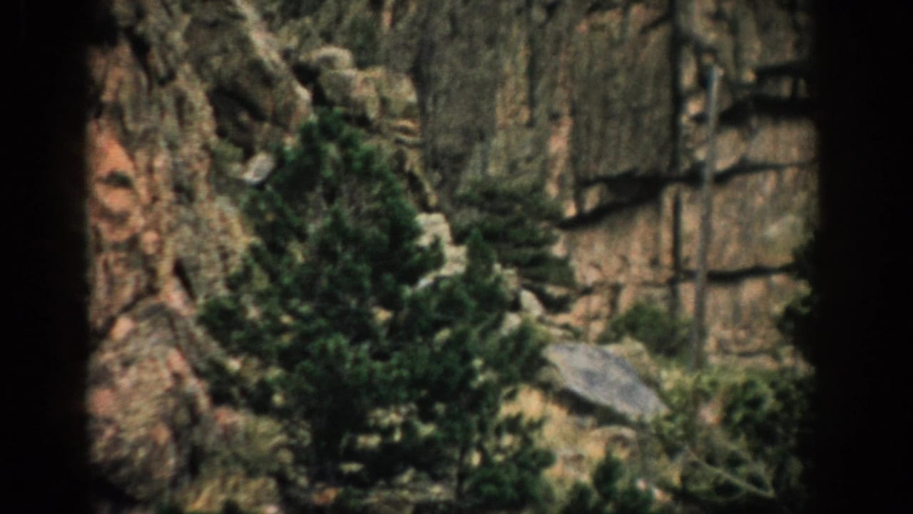 1959 Colorado home-movie frame with layered rock, scattered evergreens, shrubs, and a dark vertical border.