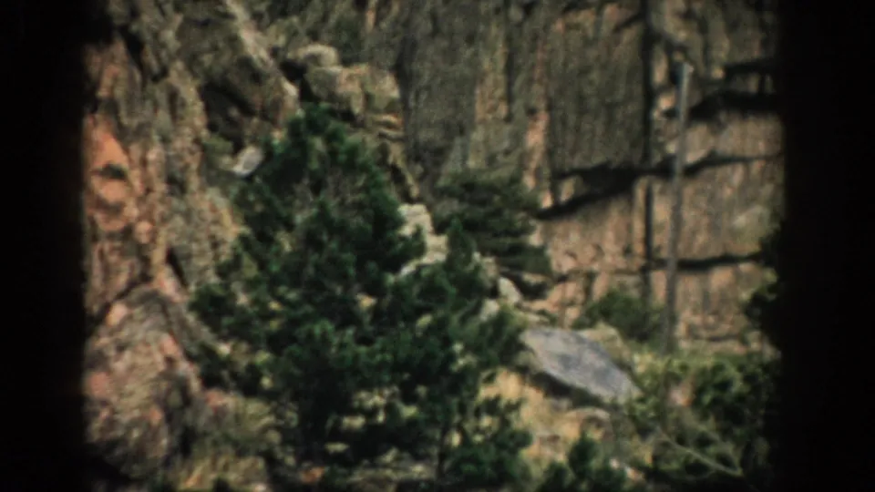 1959 Colorado home-movie frame with layered rock, scattered evergreens, shrubs, and a dark vertical border.