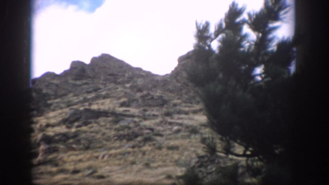 1959 Colorado frame of a rocky mountain slope with sparse grass, pale sky, and pine branches along the side.