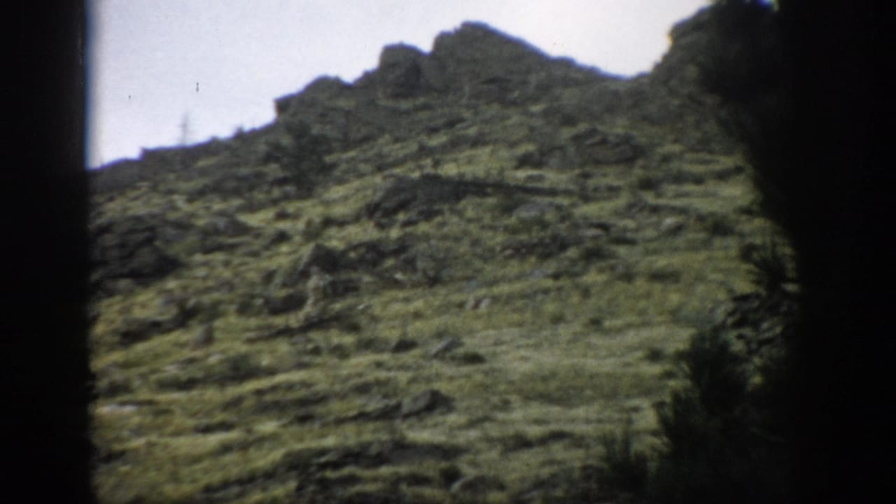 1959 Colorado 8mm frame of a rugged green slope with exposed rocks, uneven ridgeline, and dark tree silhouettes at the sides.