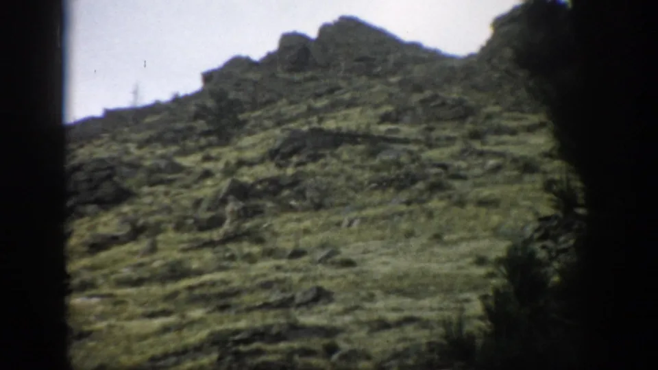 1959 Colorado 8mm frame of a rugged green slope with exposed rocks, uneven ridgeline, and dark tree silhouettes at the sides.