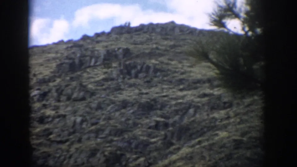 1959 Colorado 8mm frame of a rocky mountainside with low green growth, scattered trees, and a bright partly cloudy sky.