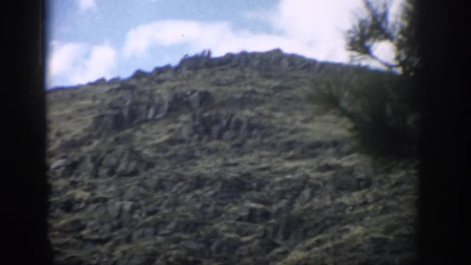 1959 Colorado 8mm frame showing rocky mountain terrain, muted green ground cover, bright clouds, and a soft tree shape near the frame edge.