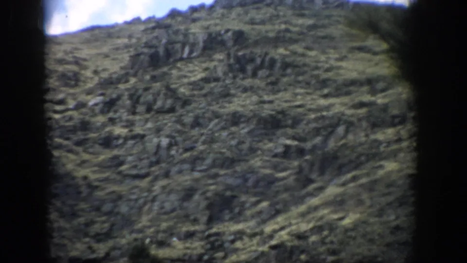 1959 Colorado 8mm frame with exposed stone ledges across a green mountainside beneath a bright, partly cloudy sky.