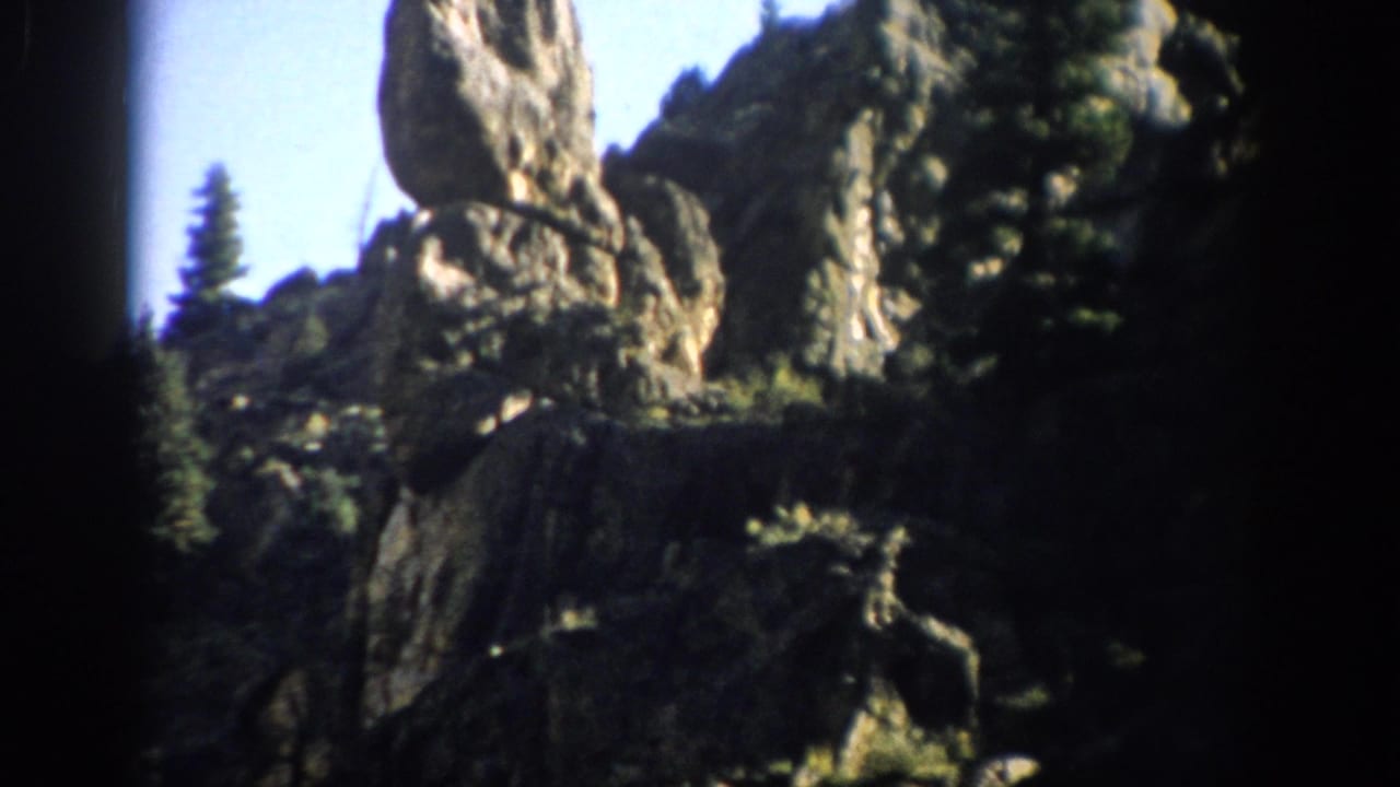 1959 Colorado 8mm frame showing rugged rock formations, dark cliff shadows, evergreen trees, and blue sky.