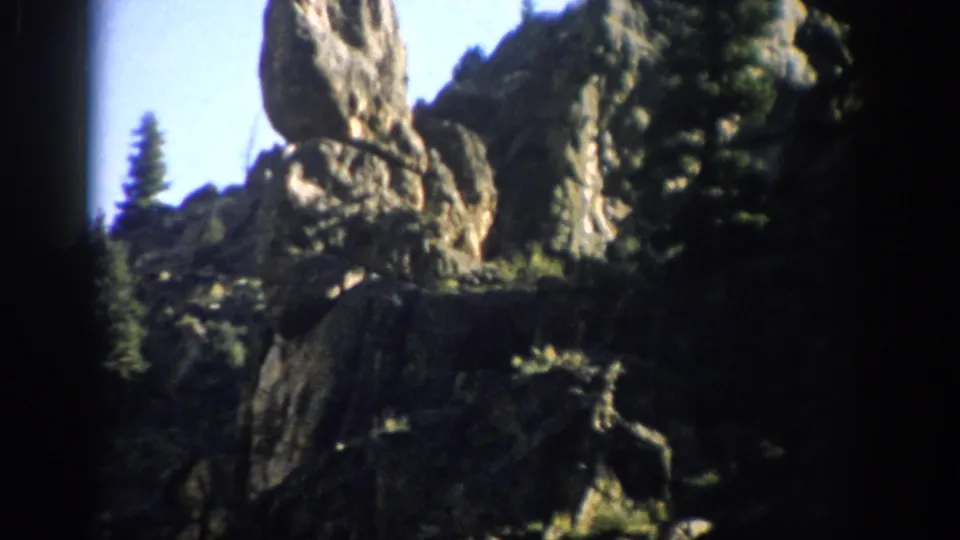 1959 Colorado 8mm frame showing rugged rock formations, dark cliff shadows, evergreen trees, and blue sky.