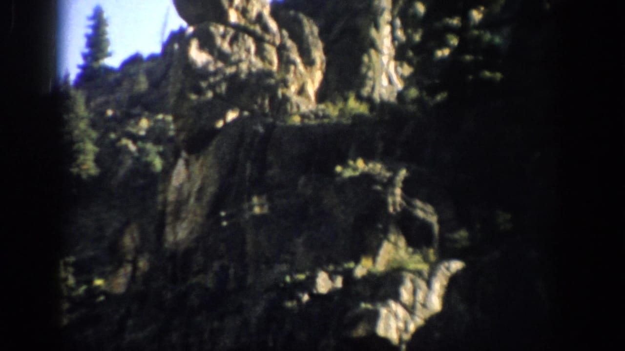 Cliff face with tall rock formation and evergreen trees under blue sky.