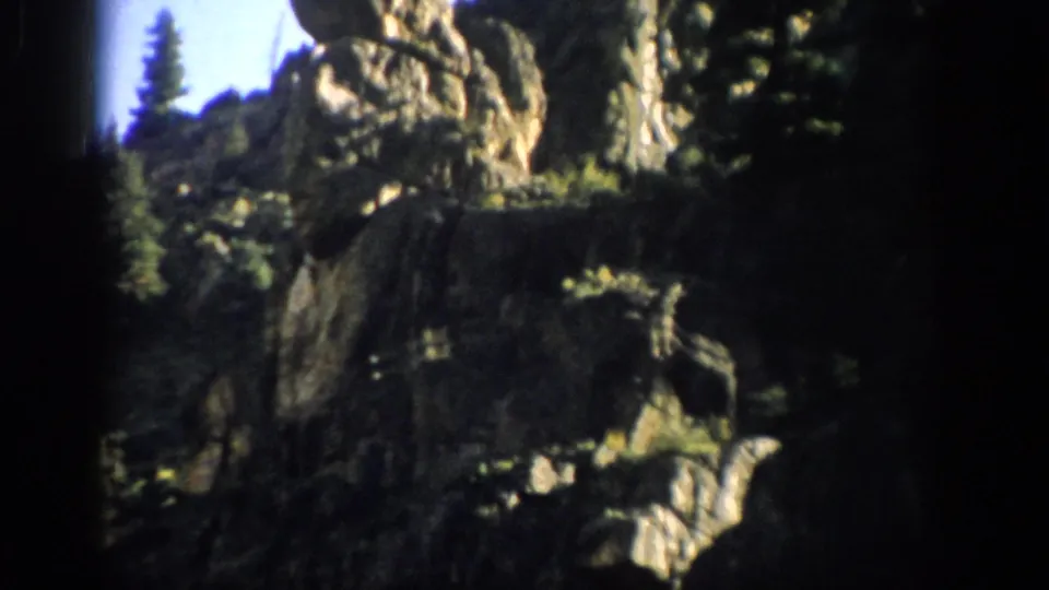 1959 Colorado home-movie frame of a tall rocky outcrop above shadowed cliffs and scattered evergreens.