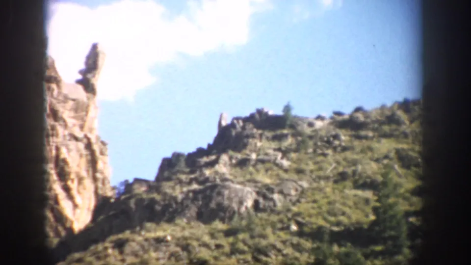 1959 Colorado 8mm view of a bright cliff edge, rocky hillside, sparse evergreens, and overexposed sky.