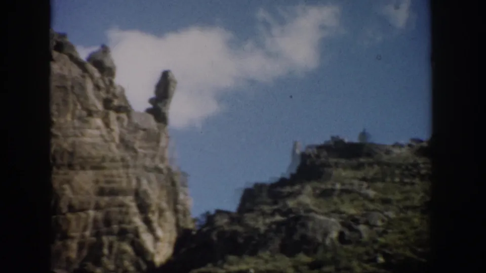 1959 Colorado frame showing a layered cliff, narrow rock spire, lower rocky ridge, and blue sky with clouds.