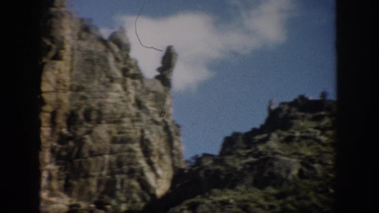 1959 Colorado 8mm frame showing a rugged cliff face, dark rock shadows, sparse greenery, and blue sky with a small white cloud.