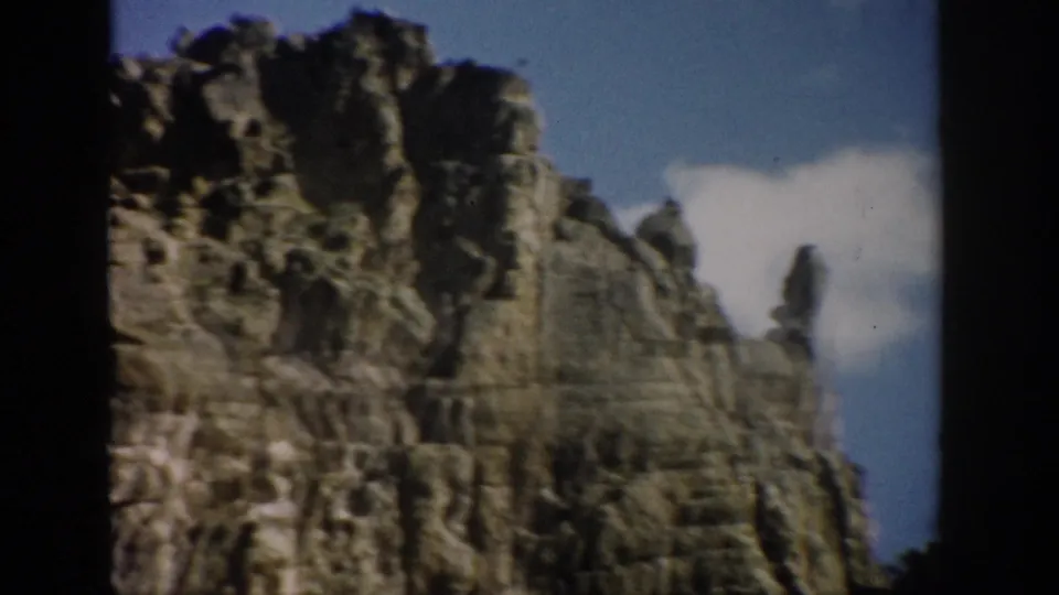 1959 Colorado home-movie frame with a layered stone formation, rough cliff crown, blue sky, and scattered white cloud.