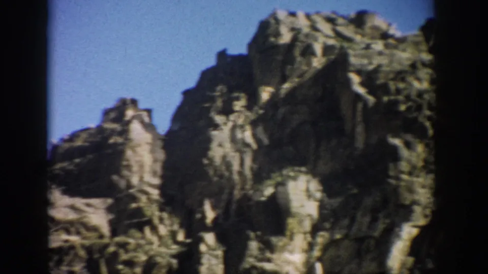 1959 Colorado frame showing a tall rugged rock formation, angular stone ledges, deep shadows, and clear blue sky.