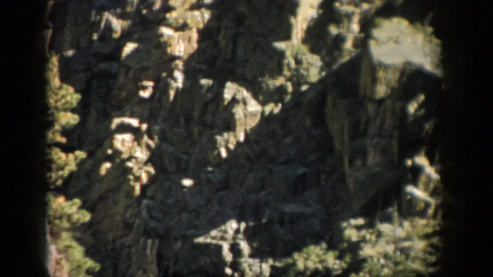 1959 Colorado home-movie frame showing shadowed stone formations and green shrubs on a rugged hillside.