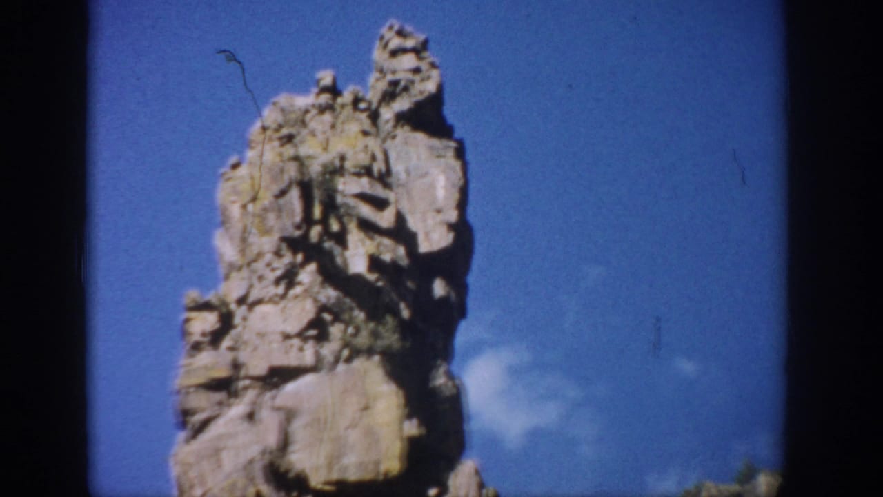 Colorado 1959 home-movie frame showing a jagged eroded rock pillar rising against blue sky with sparse brush near the base.