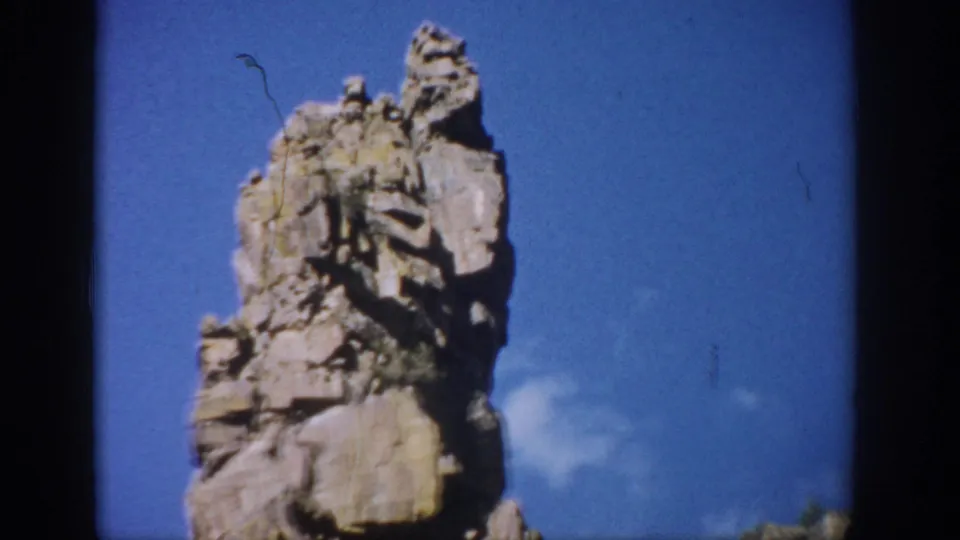 Colorado 1959 home-movie frame showing a jagged eroded rock pillar rising against blue sky with sparse brush near the base.