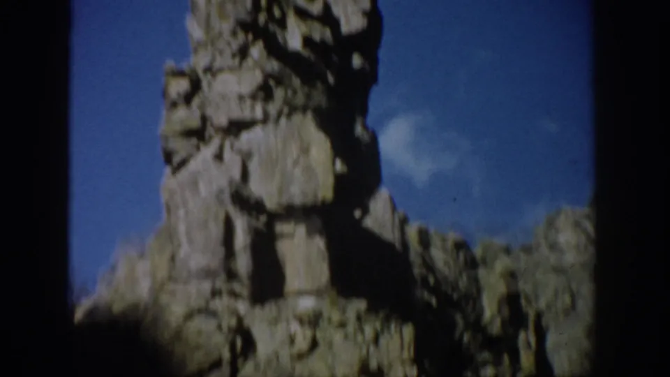 Handheld Colorado 1959 frame showing the eroded formation from a lower angle, with shadowed rock base and blue sky in the background.