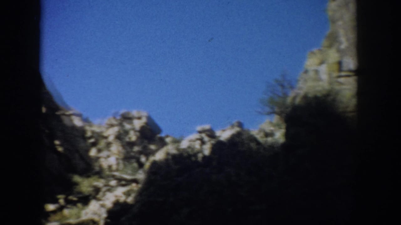 1959 Colorado 8mm frame showing jagged cliff ridges, scrub vegetation, blue sky, and heavy dark film-edge shadow.