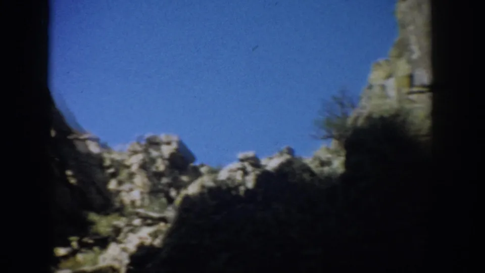 1959 Colorado 8mm frame showing jagged cliff ridges, scrub vegetation, blue sky, and heavy dark film-edge shadow.