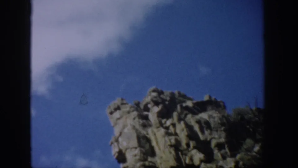 1959 Colorado 8mm view of cliff rocks from below, with blue sky, light cloud, shadowed edges, and grainy color film texture.