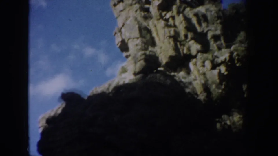 1959 Colorado frame showing a large shadowed cliff face, rugged stone texture, blue sky, wispy cloud, and dark 8mm border.