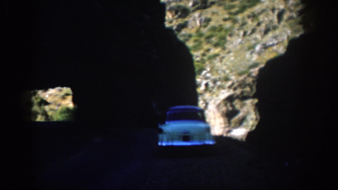 1959 Colorado home-movie frame showing a white car in deep tunnel shadow with rocky sunlit mountain terrain beyond.