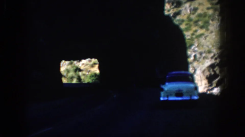 White mid-century car seen in a dark Colorado mountain tunnel, with bright rocky slopes visible through tunnel gaps.