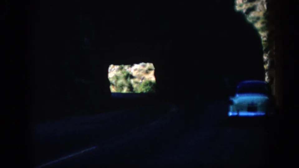 1959 Colorado road-travel frame with a white car on a shadowed tunnel road and sunlit rock visible through an opening.