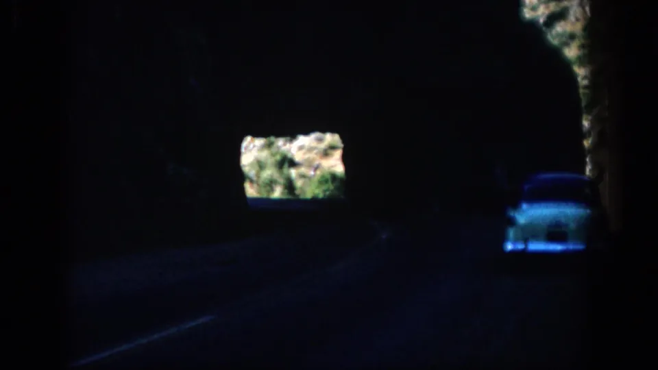 Dark 8mm frame from Colorado showing a white car inside a mountain tunnel with sunlit rocky terrain beyond.