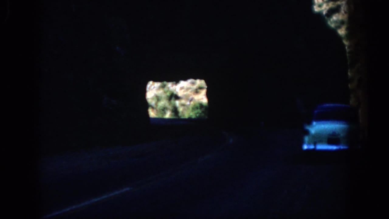 White car seen ahead inside a dark rock tunnel with a bright opening and foliage beyond.