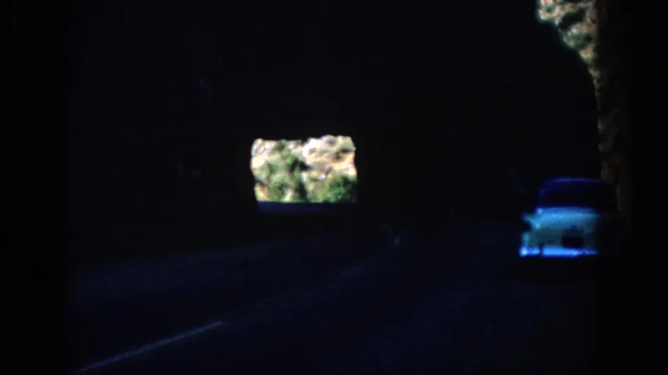 1959 Colorado 8mm frame showing a white car driving through a shadowed rock tunnel toward a sunlit opening.