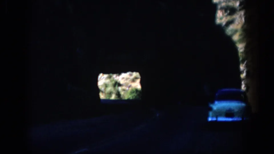 White car continues through a dim Colorado rock tunnel, with daylight and rough stone visible at the exit.