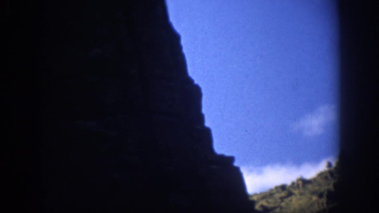 1959 Colorado home-movie frame showing a dark rock face in silhouette, blue sky, and a sparse sunlit canyon slope.