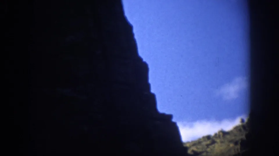 1959 Colorado home-movie frame showing a dark rock face in silhouette, blue sky, and a sparse sunlit canyon slope.