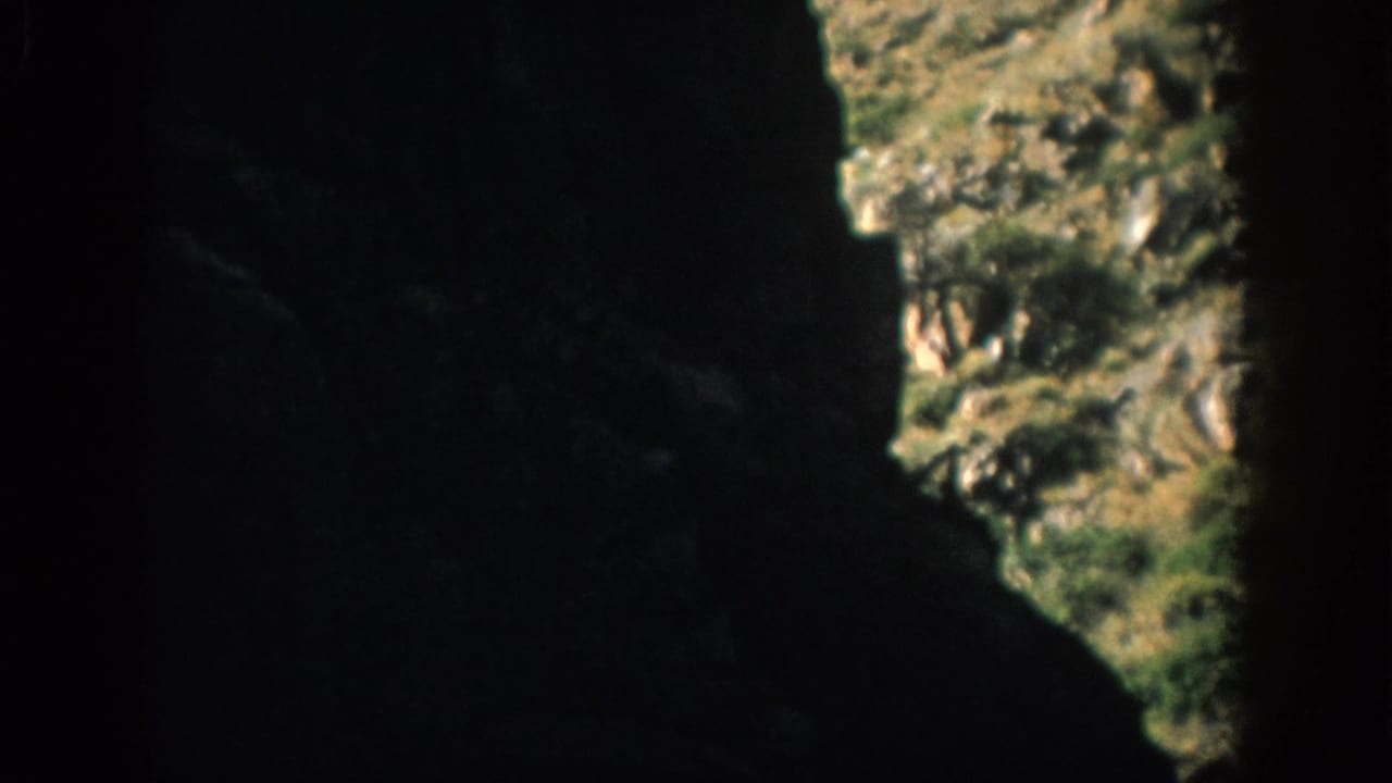 Dark cave or tunnel interior with a bright irregular opening onto grass and rock.