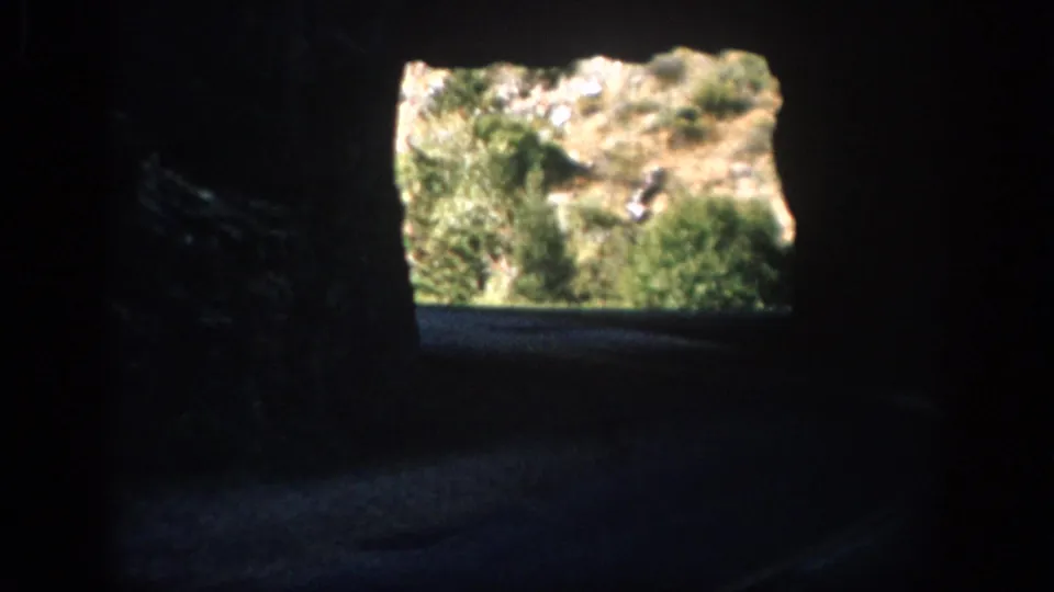 1959 Colorado home-movie frame from inside a dark road tunnel, looking toward a sunlit hillside and vegetation at the exit.