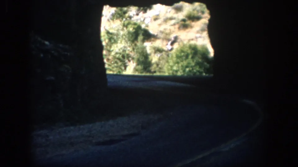Inside a dark Colorado road tunnel, the road curves toward a bright exit with rocky brush-covered terrain beyond.