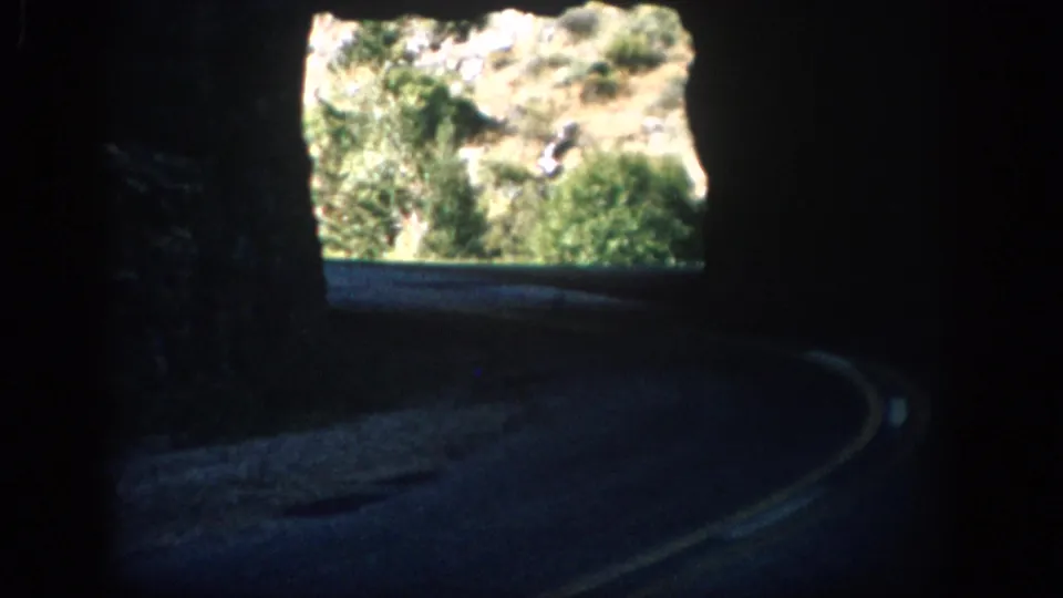 A low-light 8mm frame shows the vehicle view through a tunnel toward sunlit Colorado hillside vegetation.