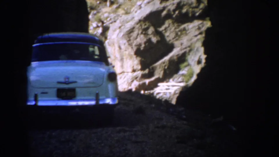The blue mid-century sedan exits the tunnel into bright Colorado mountain terrain, framed by deep shadow and rock.