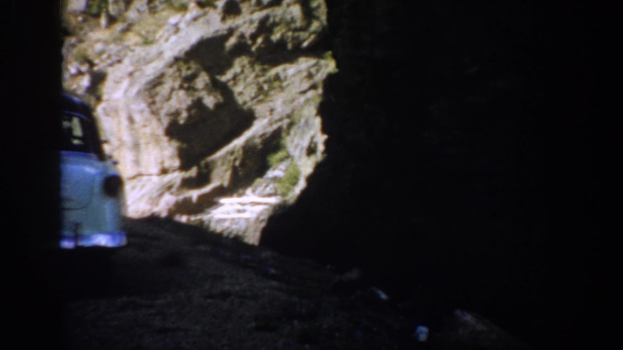 1959 Colorado home-movie frame showing a pale sedan edge near a dark cave or tunnel exit with rocky cliff in bright daylight.